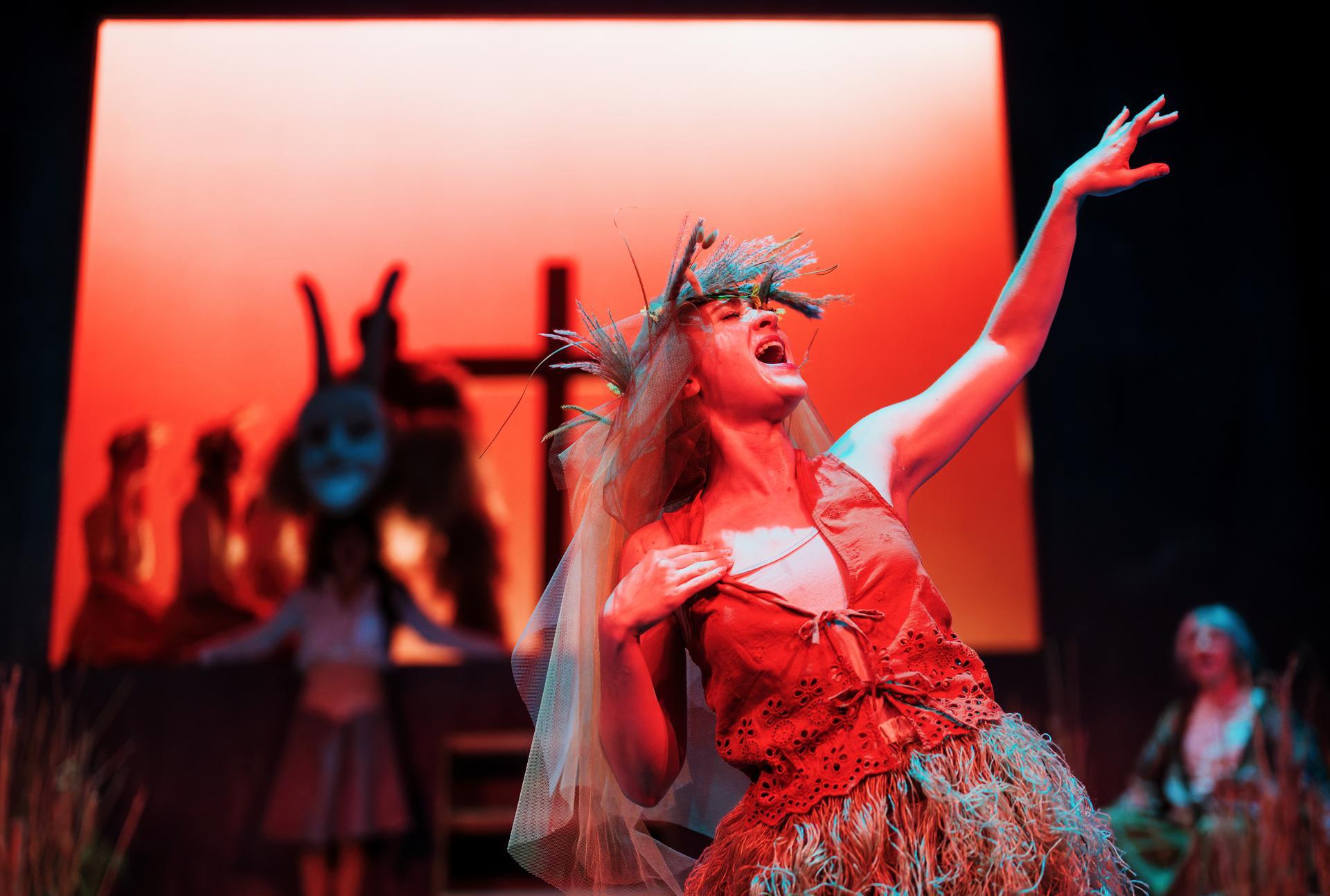 Performer in a red costume and feathered headdress reaches upwards mid-cry on stage, with masked figures and a cross-like structure in the red-lit background